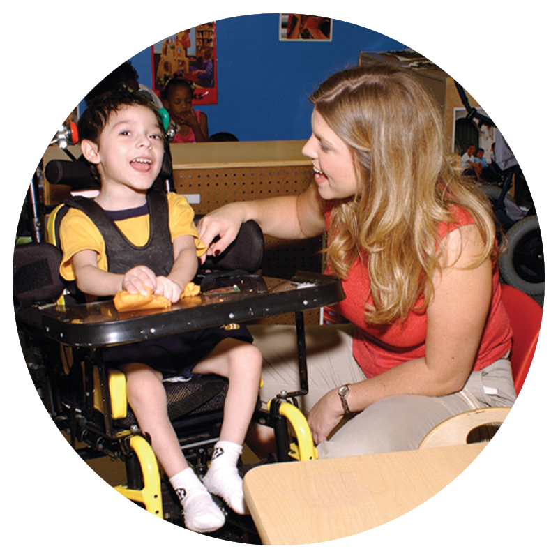 A teacher sits smiling beside a young student in a mobility device