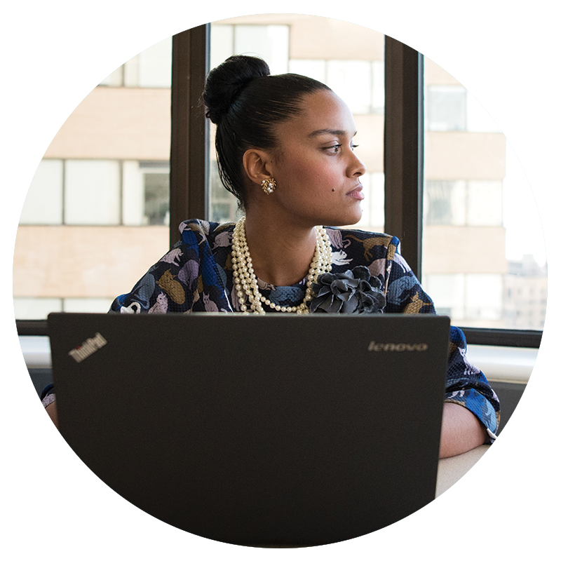 circular cropped image of a young professional woman of color seated in front of a laptop, looking off to the side