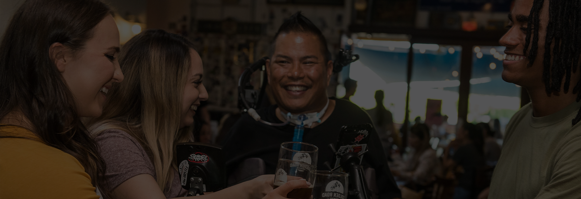 A group of four friends, including a man using a power wheelchair with assistive technology and a tube, are gathered at a lively indoor venue, smiling and toasting with drinks.